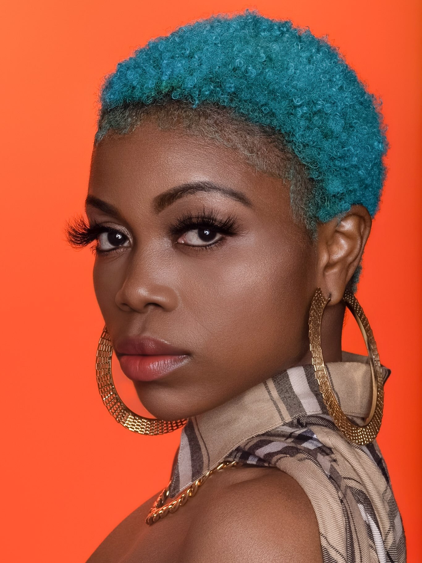Editorial portrait with a teal curly crop and gold hoops against a burnt-orange backdrop